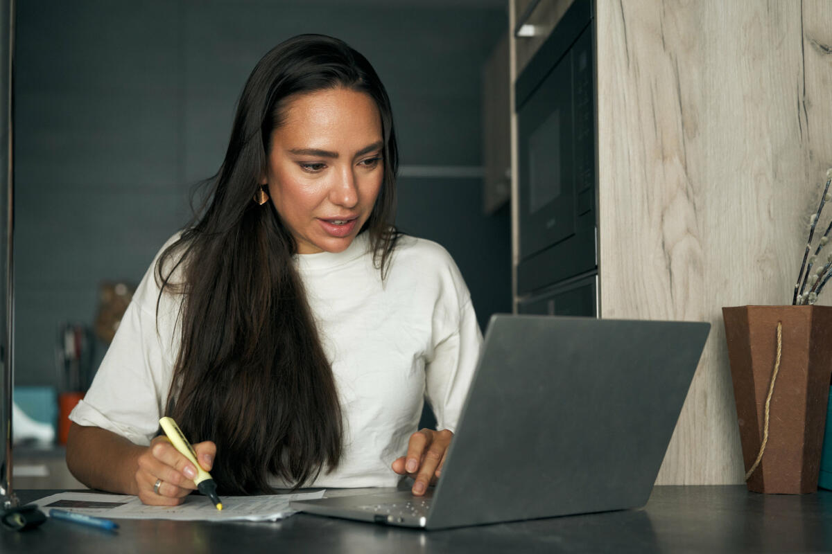 Frau sitzt am Schreibtisch mit Laptop und markiert etwas auf dem Zettel © Sergey Mironov / GETTY IMAGES