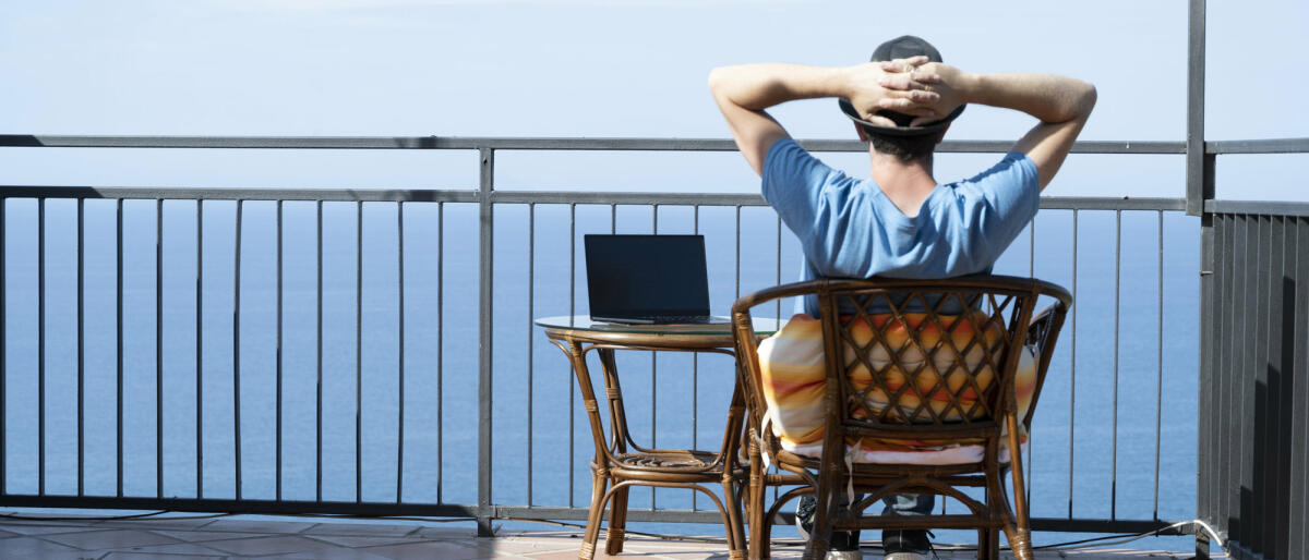 Mann sitzt mit Laptop auf Terrasse und blickt auf Meer © Marco Bottigelli / Getty Images