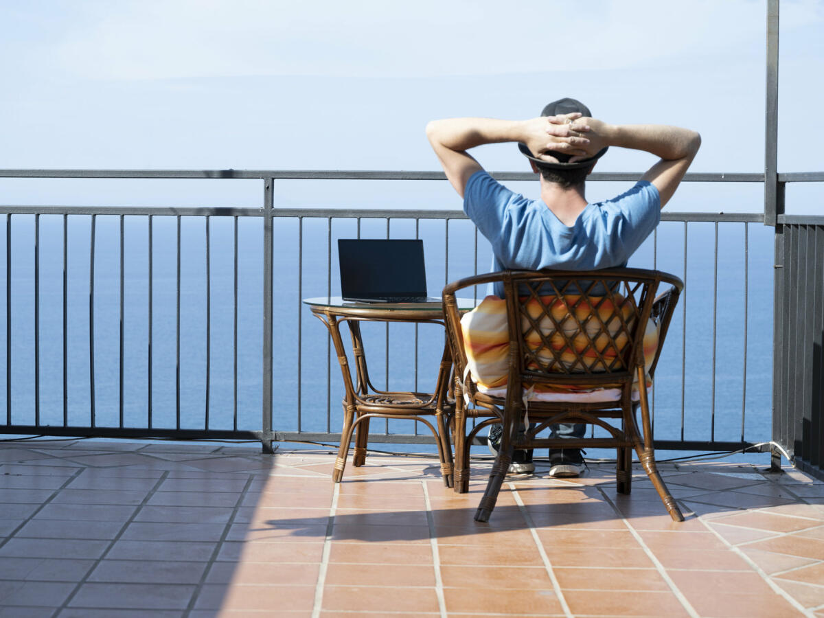Mann sitzt mit Laptop auf Terrasse und blickt auf Meer © Marco Bottigelli / Getty Images